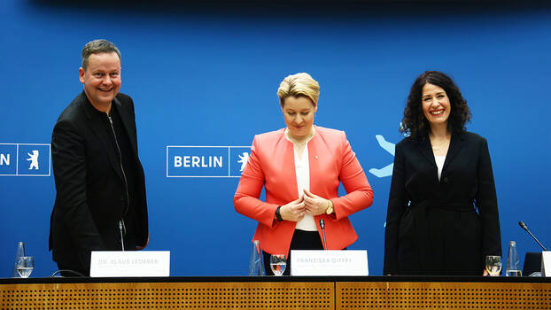 Franziska Giffey, Bettina Jarasch, and Klaus Lederer stand in front of a podest during a press conference.