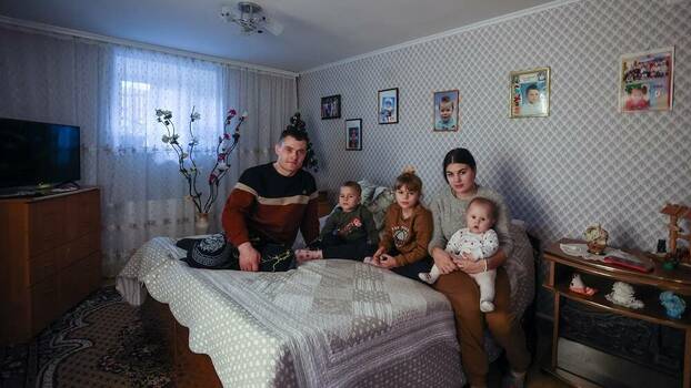 Two adults and three children sit on a bed in front of family pictures.
