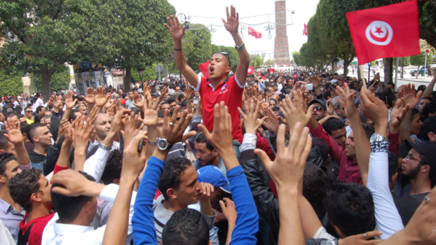Menschenmasse, eine Demo. Rechts im Bild ist die tunesische Flagge. Ein Mann sticht hervor, er scheint etwas zu schreiben, seine Arme sind in der Luft.