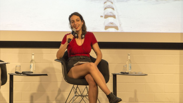 [Translate to en:] Karina Nohales über den Prozess der verfassungsgebenden Versammlung in Chile auf dem Festival de:lux im Juni 2022. Sie sitzt auf einem Stuhl auf einer Bühne.