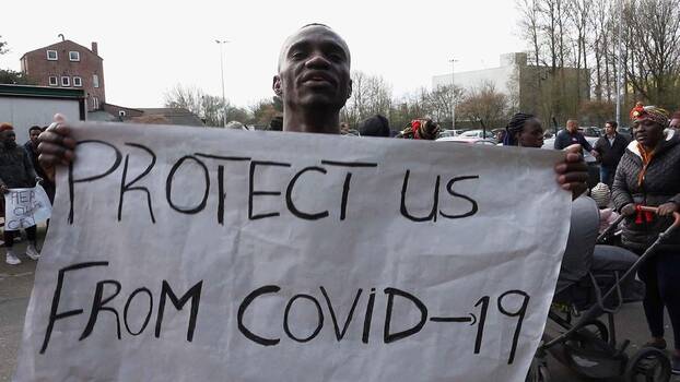 Ein junger Mann hält ein Bettlaken nach oben. Auf dem laken steht: «Protect us from Covid-19 (Schützt uns vor Covid-19)»
