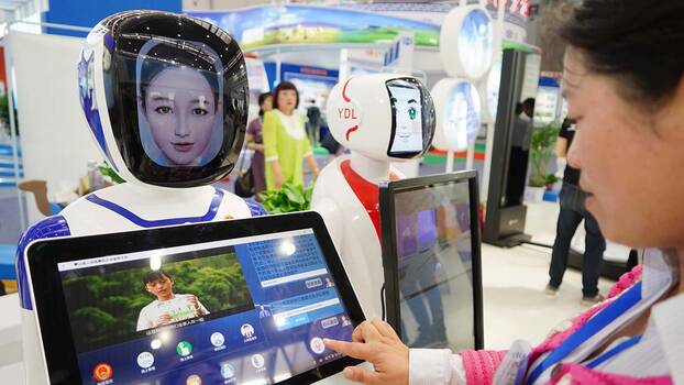 [Translate to en:] Eine Frau steht in einer Messehalle vor einem Monitor, auf dem sie mit der Hand etwas auswählt. Der Monitor wird von einer Roboterfigur gehalten, die die Frau mit einem künstlichen aber menschlich wirkendem Gesicht anlächelt. 