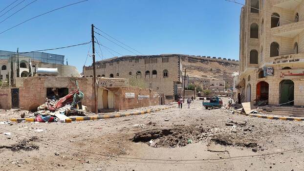 Ein Straßenzug in der nach einem Luftwaffenangriff der von Saudi-Arabien geführten Koalition im April 2015 zerstörten Hauptstadt Jemens, Sana'a. In der Mitte der unasphaltierten Straße ist ein großes Loch im Schotter. Rechts der Rohbau eines ehemaligen Hauses, es fehlen sämtliche Fensterscheiben.  fenster
