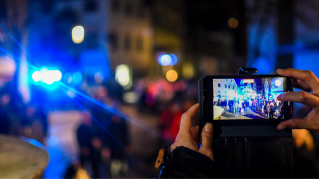 Ein Smartphone wird von einer Hand gehalten, auf dem Bildschirm sind Demonstranten und Lichter zu erkennen, die im Hintergrund des Bildes ebenfalls undeutlich zu sehen sind. 