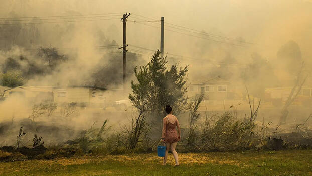Eine Person im kurzen Sommerkleid, mit einem blauen Wassereimer in der Hand, läuft barfuss über eine Wiese. Im Hintergrund ist durch dichte Rauchwolken Stromleitungen und ein Haus zu sehen.