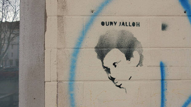 [Translate to en:] Ein Graffiti mit dem Konterfei von Oury Jalloh, ist in shcwarzer Farbe an eine Mauer gesprüht. Aufgenommen in Neukölln, Berlin im Dezember 2017. Unbekannte*r Künstler*in.