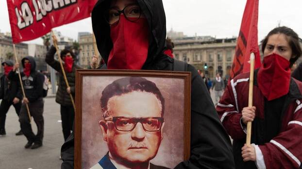 Protesters march carrying red flags and a portrait of Salvador Allende.