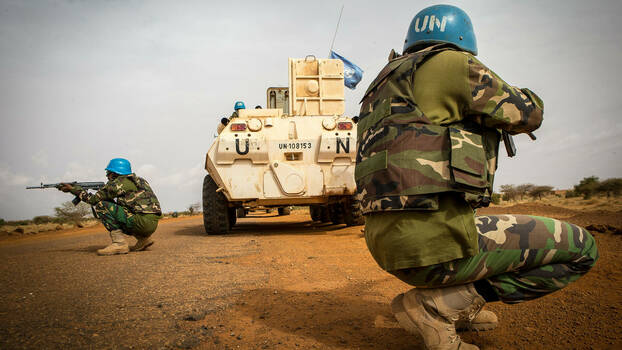 Zwei Soldaten mit blauen UN-Helmen knien mit Gewehren im Anschlag hinter einem UN-Panzerfahrzeug im Sand.