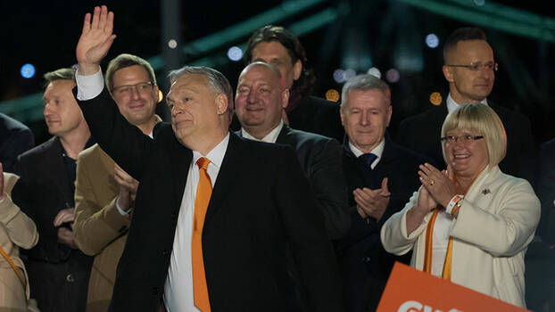Viktor Orban steht im Vordergrund und winkt. Hinter ihm viele Leute, die klatschen.