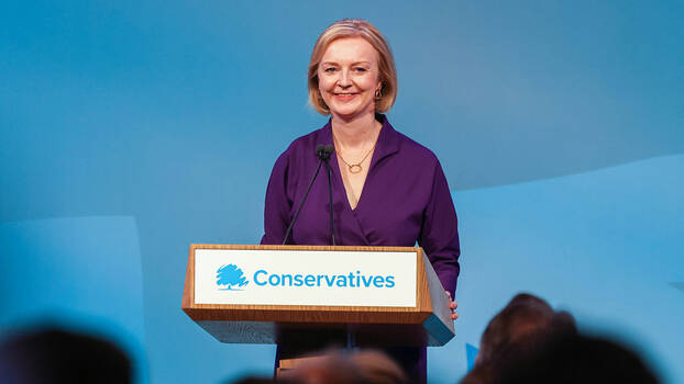 Liz Truss mit lila Oberteil an einem Redepult auf dem "Conservatives" steht.