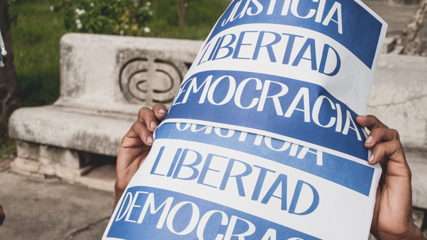 Aufnahme von einer Demonstration im Juli 2018 in Granada, Nicaragua. Zwei Hände halten ein Plakat mit der Aufschrift «Justicia, Libertad, Democracia (Gerechtigkeit, Freiheit, Demokratie)» Bild: Julio Vannini, CC BY-NC 2.0