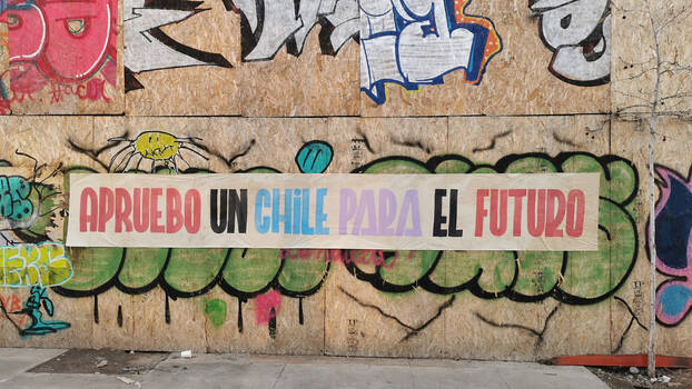 [Translate to en:] Ein Banner an einer Wand mit Graffiti wirbt für die neue Verfassung. «Apruebo un Chile para el futuro» - Zustimmung für ein Chile der Zukunft - steht dort in großen bunten Buchstaben. 