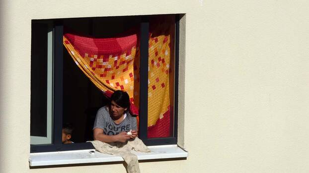 Eine Frau schaut aus dem Fenster ihres Zimmers in einer Unterkunft für Geflüchtete. Als Vorhang hat sie ein gelb-rotes Tuch angebracht.. 