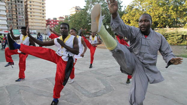 A group of Black men shows kungfu on a public place. Most men wear red pants and a white shirt, one man in the front wears grey. The clothes look Chines. All do a kick with their right leg.