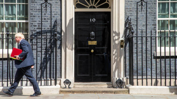 Boris Johnson geht mit großem Schritt und roter Mappe unter dem Arm auf die linke Bildkante zu. Hinter ihm der Eingang von 10 Downing Street.