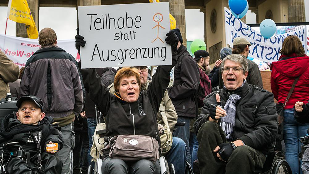 Zu sehen ist der Ausschnitt einer Demo tagsüber und draußen vor dem Brandenburger Tor in Berlin. Im Vordergrund sind 3 Personen im Rollstuhl. Die Person in der Mitte hält ein Pappschild hoch. Darauf steht: „Teilhabe statt Ausgrenzung.“ Daneben ist eine orangene Strichfigur gezeichnet die ein lachendes Gesicht hat. Die Person rechts im Bild hält einen Daumen hoch. Die 3 Personen haben helle Haut und sind ca. zwischen 40-65 Jahre alt. Im Hintergrund sind weitere Personen, Transparente und Banner zu sehen. Die Personen tragen herbstliche Kleidung. 