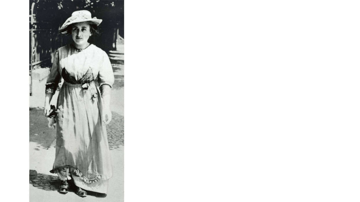 Zu sehen ist eine schwarz-weiß Fotografie von Rosa Luxemburg tagsüber und draußen.  Sie läuft eine Straße entlang. Sie trägt ein weißes Sommerkleid und einen weißen Hut. Sie hat helle Haut.